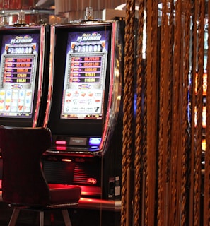 A pair of casino slot machines, each with detailed screens displaying various game outcomes and jackpot amounts. The chairs in front of the slot machines are dark and cushioned for comfort. The background features a hanging curtain made of golden ropes and other casino decorations are visible.