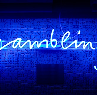 A bright blue neon sign spells out the word 'gambling' in cursive script. The background features a textured wall covered in domino tiles, also illuminated by the blue light, creating a vivid and visually striking scene.