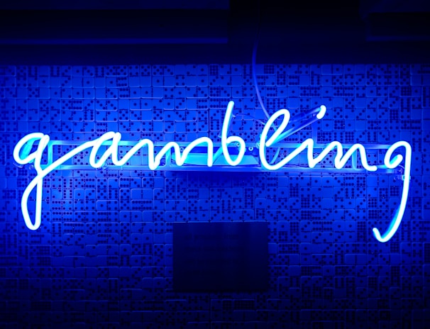 A bright blue neon sign spells out the word 'gambling' in cursive script. The background features a textured wall covered in domino tiles, also illuminated by the blue light, creating a vivid and visually striking scene.