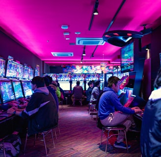 A vibrant arcade with rows of gaming machines adorned with colorful screens. Several people are immersed in playing games, seated on stools in a dimly lit room accented by an intense pink ceiling light. The atmosphere is lively and engaging.