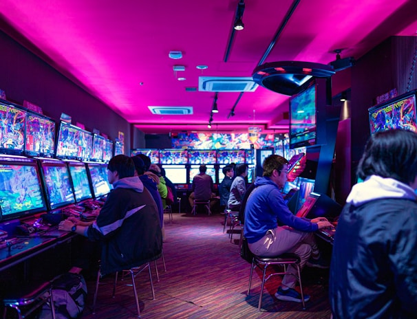 A vibrant arcade with rows of gaming machines adorned with colorful screens. Several people are immersed in playing games, seated on stools in a dimly lit room accented by an intense pink ceiling light. The atmosphere is lively and engaging.