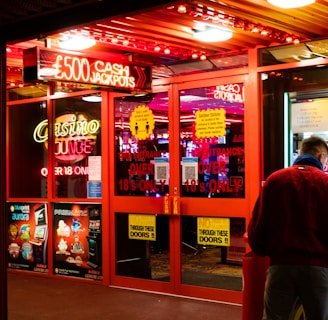 A brightly lit entrance to a casino with neon signs, including one advertising a £500 jackpot. The doors have clear signage indicating entry is restricted to those over 18. A person in a red jacket stands to the right, possibly interacting with a machine or display.