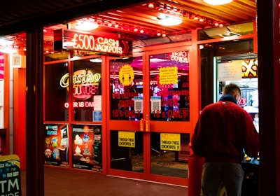 A brightly lit entrance to a casino with neon signs, including one advertising a £500 jackpot. The doors have clear signage indicating entry is restricted to those over 18. A person in a red jacket stands to the right, possibly interacting with a machine or display.