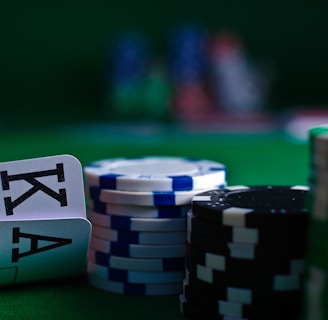 A casino scene featuring two playing cards, the ace and king of clubs, partially concealed by a player's hand. Nearby, there are stacks of multicolored poker chips on a green felt table, with blurred colorful background elements suggesting other chip stacks or cards.