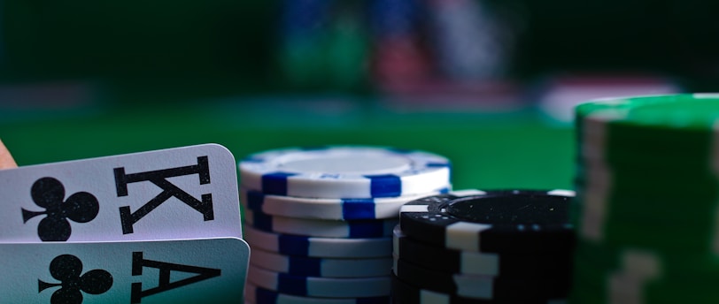 A casino scene featuring two playing cards, the ace and king of clubs, partially concealed by a player's hand. Nearby, there are stacks of multicolored poker chips on a green felt table, with blurred colorful background elements suggesting other chip stacks or cards.