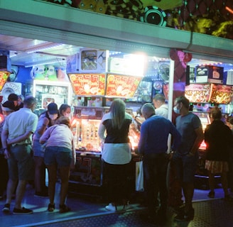 A group of people standing in front of an illuminated amusement arcade, engaging with various gaming machines. The environment is bustling with activity, and colorful lights create a vibrant atmosphere. The ceiling is decorated with large, playful images of dice and other game-related graphics, enhancing the lively mood.