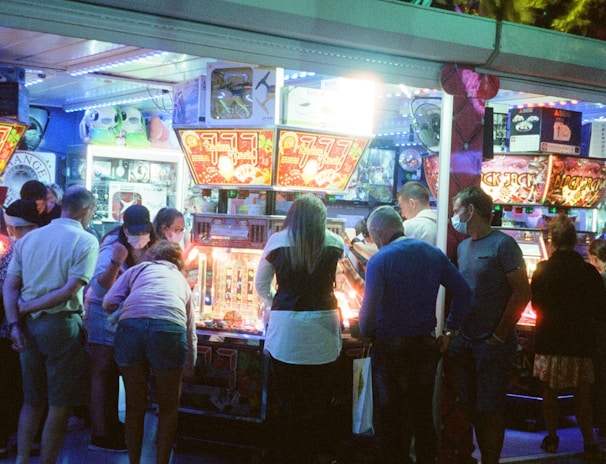 A group of people standing in front of an illuminated amusement arcade, engaging with various gaming machines. The environment is bustling with activity, and colorful lights create a vibrant atmosphere. The ceiling is decorated with large, playful images of dice and other game-related graphics, enhancing the lively mood.