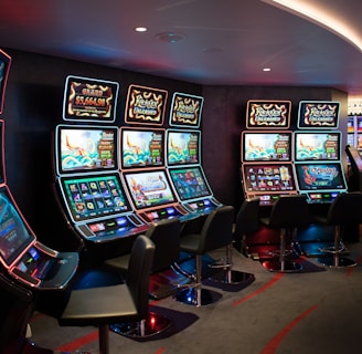 A row of brightly lit slot machines in a casino setting, each with colorful displays and various game themes. The machines are lined up against a dark wall and are unoccupied, with cushioned chairs placed in front of them. The area is dimly lit except for the glow from the screens, creating a vibrant and engaging atmosphere.
