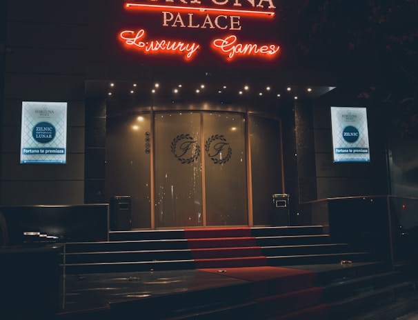 A neon-lit casino entrance with a red carpet leading up to large double doors. The sign reads 'Fortuna Palace Luxury Games' in bright red and white neon lights. The dimly lit atmosphere suggests a night-time setting. There are two digital displays on either side of the entrance showing promotional content.